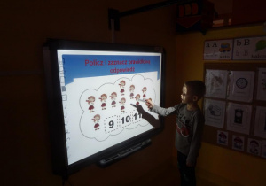 Chłopiec stoi pod tablicą interaktywną przelicza ilość dziewczynek na obrazku.